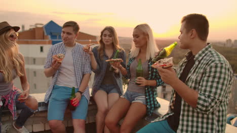 Young-frieds-at-sunset-on-the-roof-enjoy-pizza-and-beer.-They-sit-and-talk-to-each-other.